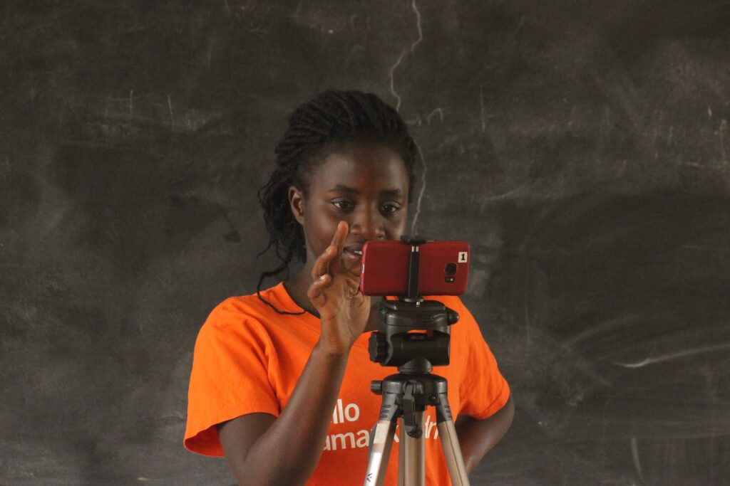 A young woman filming a scene using a smart phone. She wears an orange shirt and stands in front of a blackboard.