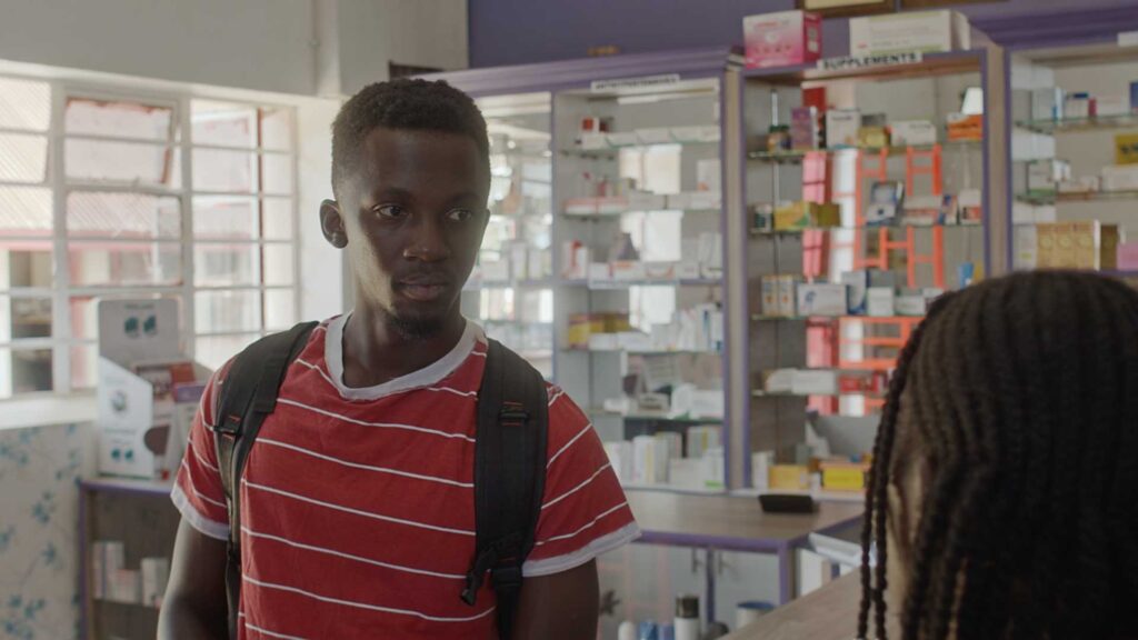 A screengrab of a film scene in a pharmacy. Medium shot of a young black actor in a red shirt with thin white stripes in dialogue with an actress on the left of the screen, only back of her head seen, she is out of focus. A large window and shop shelves full of medicines are in the background