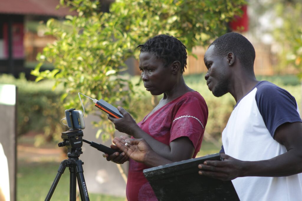 A woman stands behind a phone mounted on a tripod, while a man watches. They are filming a scene in a film class.