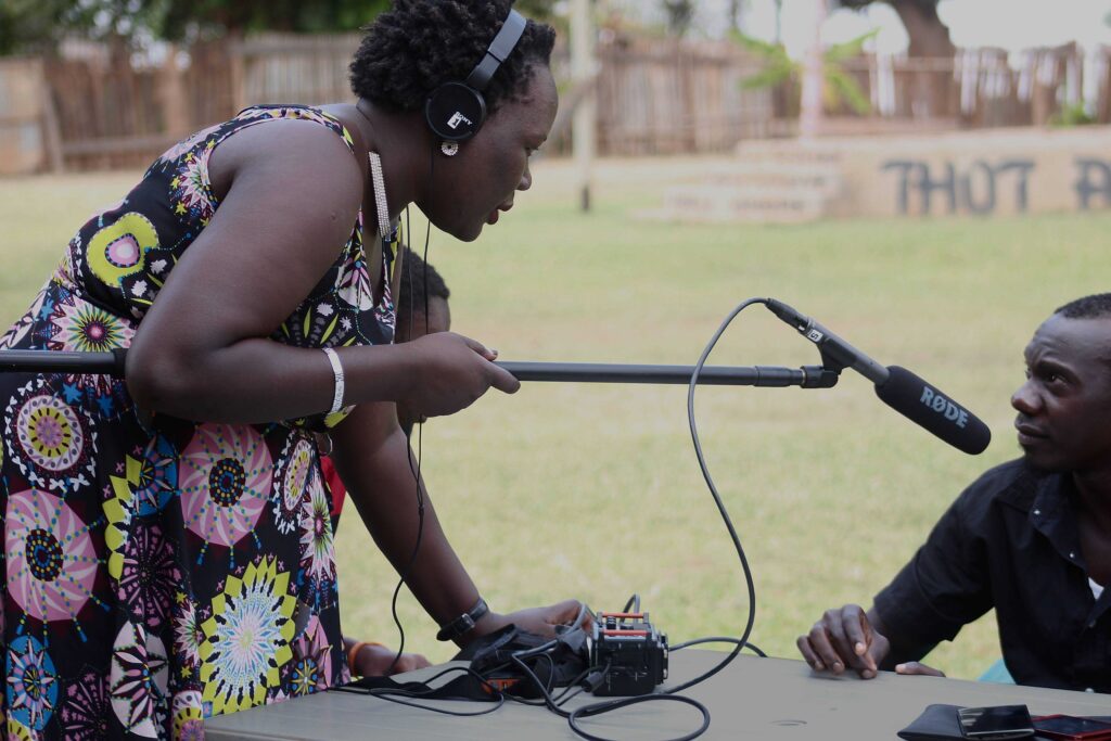A woman leans over a table on which is an audio recorder. She holds a boompole with a boom micrphone pointed at a man sitting on the other end of the table.