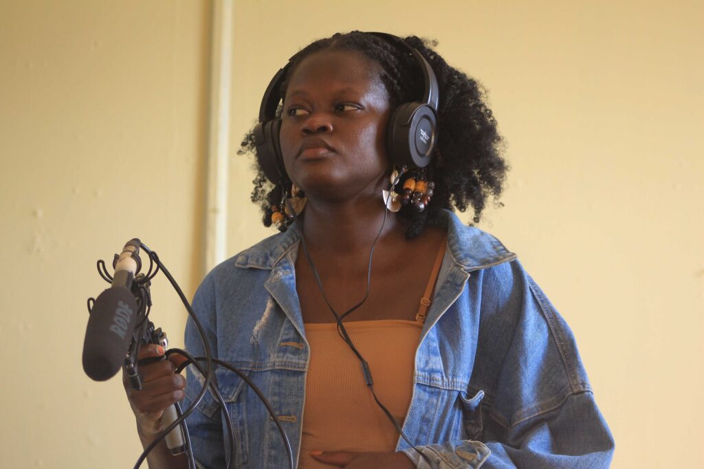 A young African woman wearing headphones and holding a boompole and a shotgun microphone.