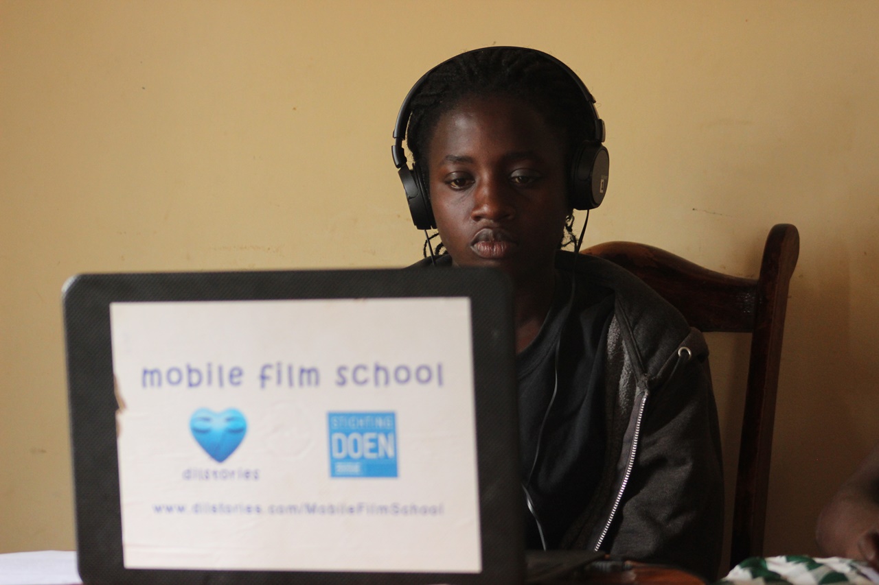A young African woman sits behind a laptop, which has a sticker with two blue logos on it, apparently working on a film editing project