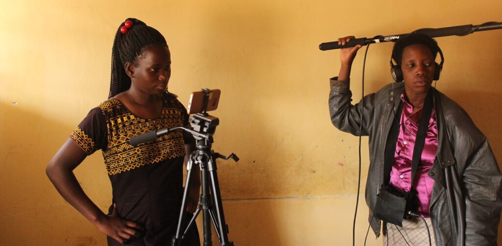 A photo of filmmakers shooting a film, a woman using a smart phone on a camera tripod, while another holds a boom. 