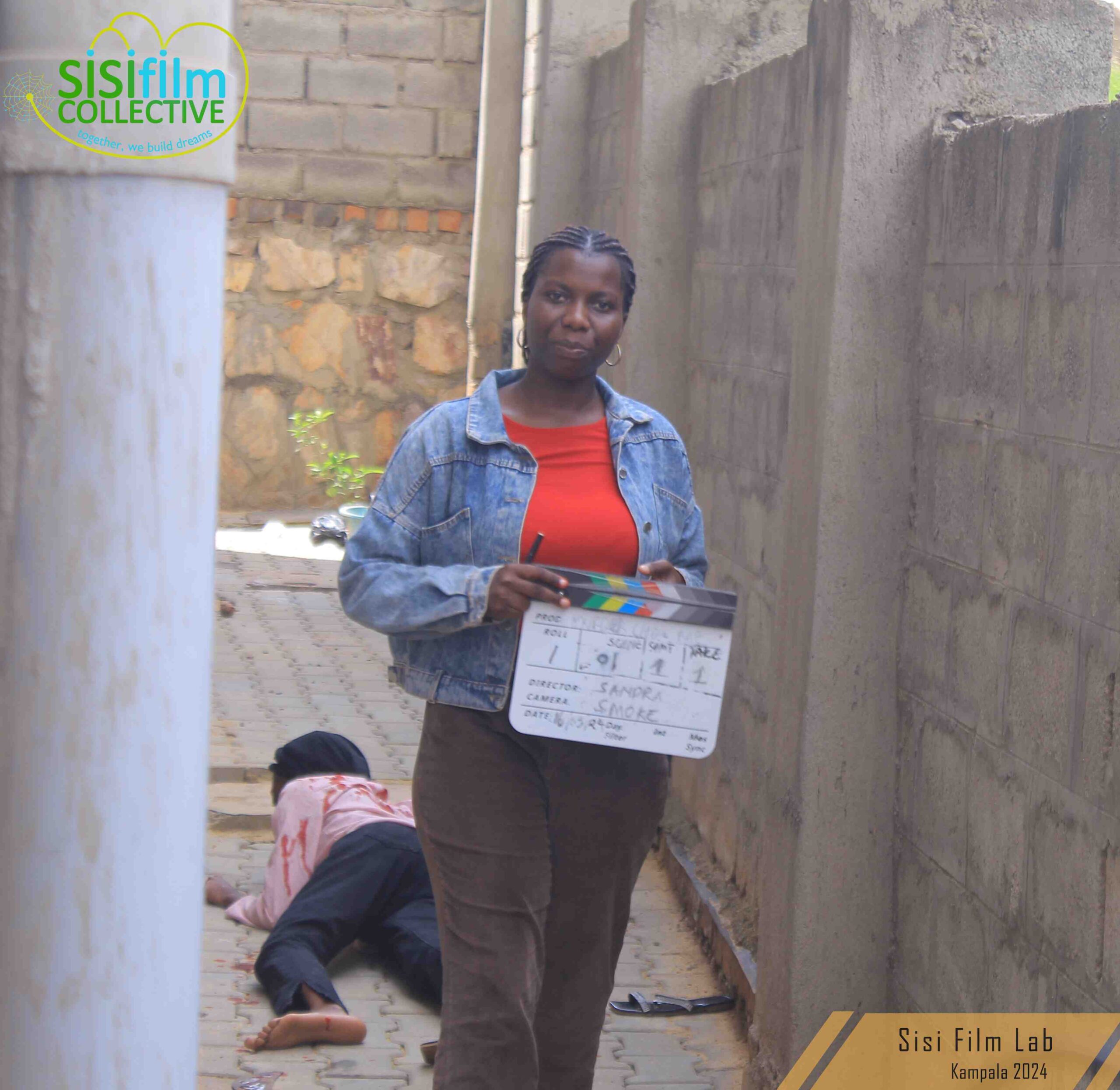 A black African woman dressed in a red blouse and a denim jacket is holding a slate. a dead body of a woman lies behind her.