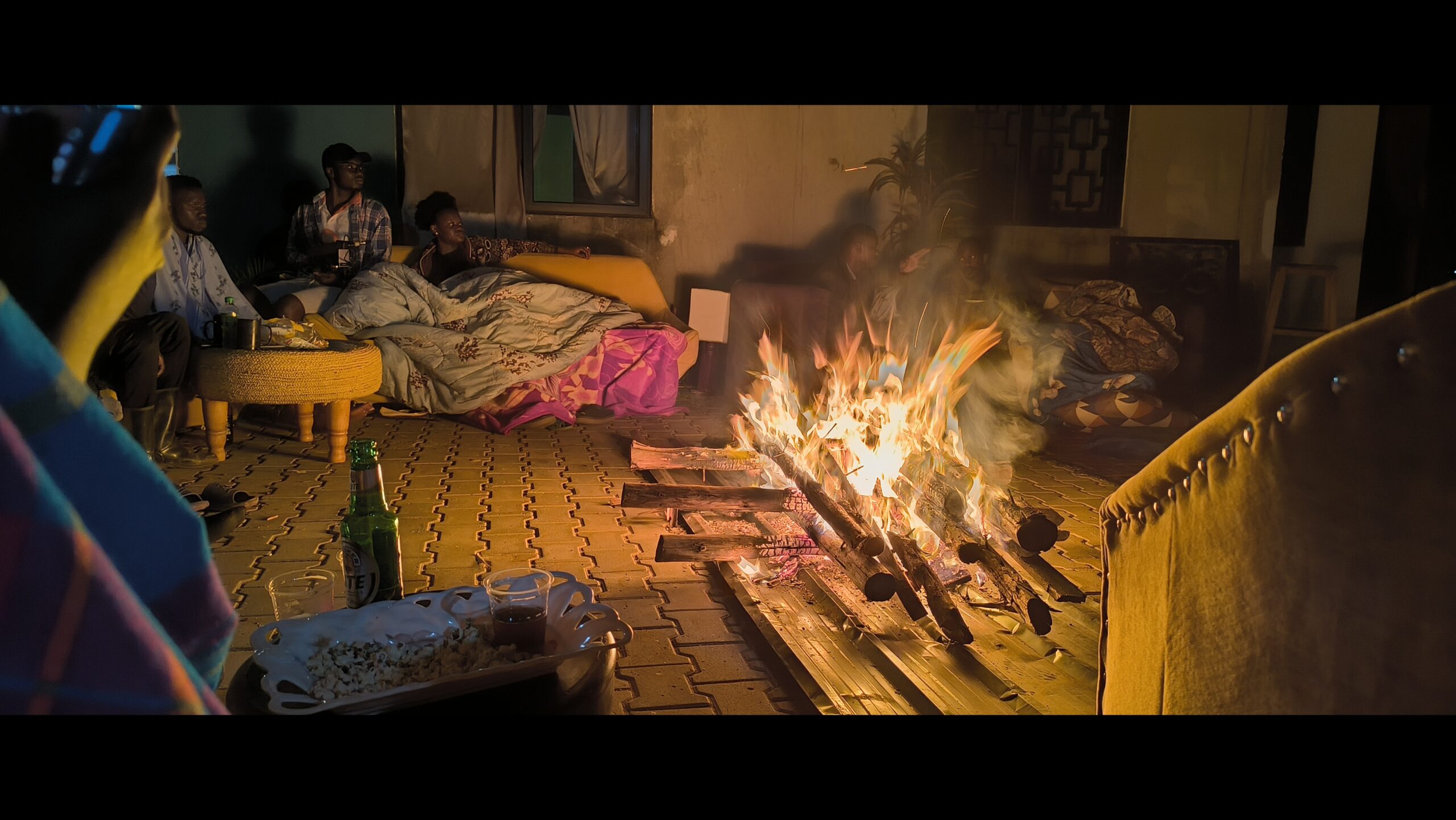 A group of people sitting together in a compound delighting in each other's presence while gathered around a bonfire to watch a movie in an outdoor film screen.