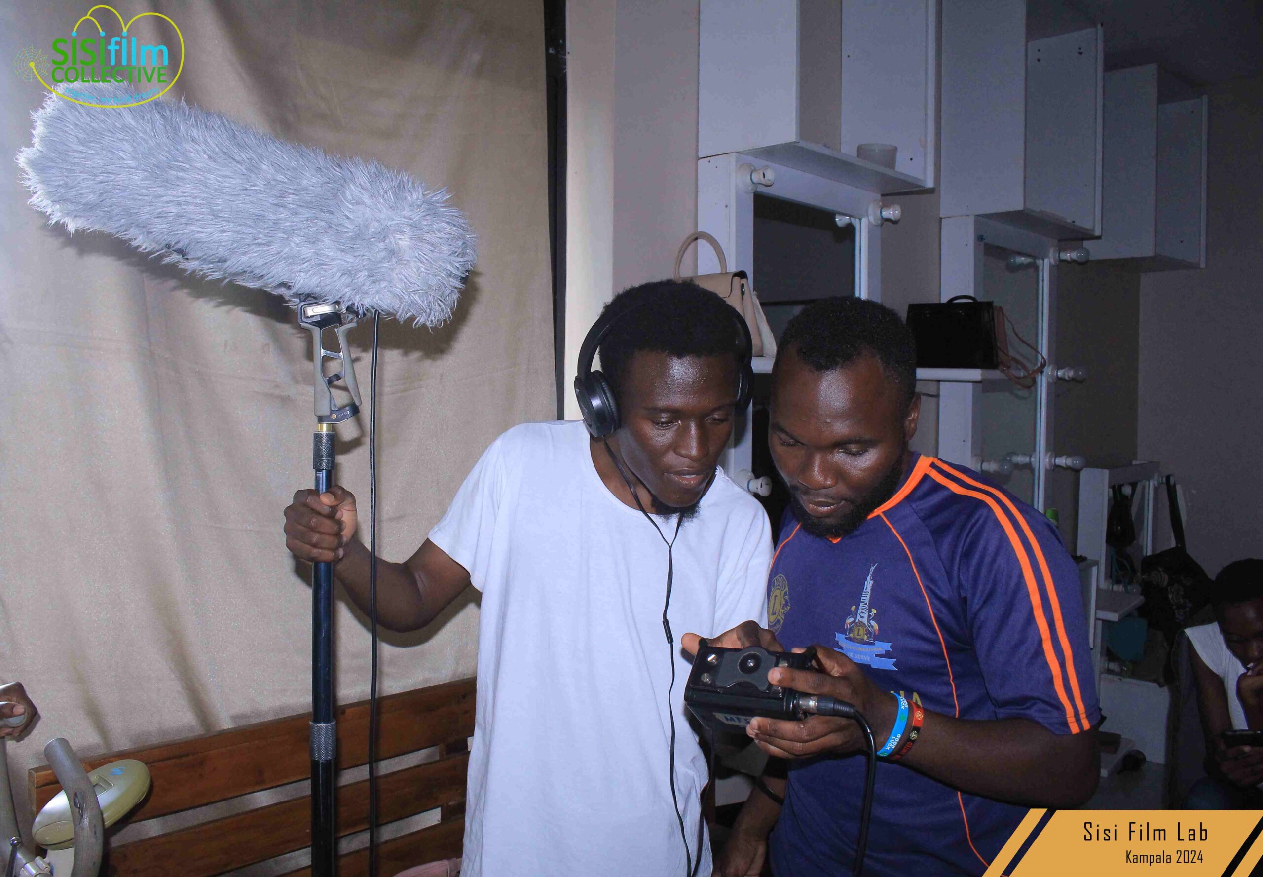 Two Black African boys, one dressed in white holding a boom with a blimp on it, he is wearing headphones staring down at a Tascam recorder being held by the other boy wearing a blue jersey with orange strips.