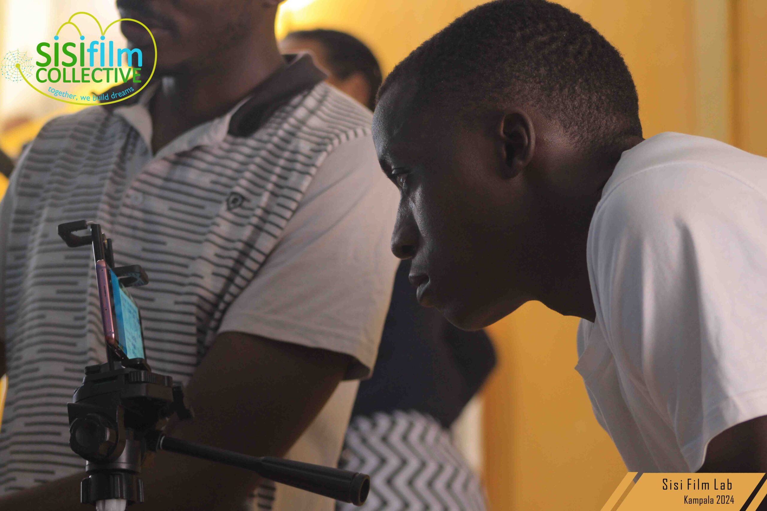 A Black African man dressed in white is bent staring at a phone standing on a tripod. He's head and shoulders are the only visible body parts in the camera. a body of a man standing in the background.