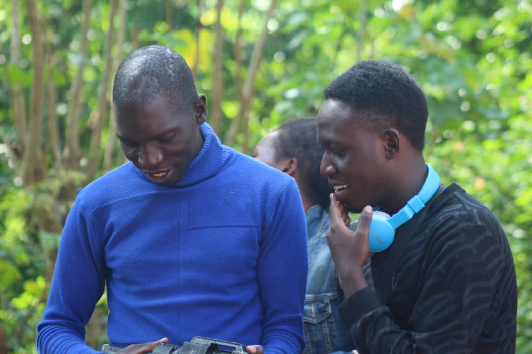 A Black African man dressed in blue is smiling while staring at something in his hands. besides him is a man smiling staring down too dressed on a black sweater with blue headphones.