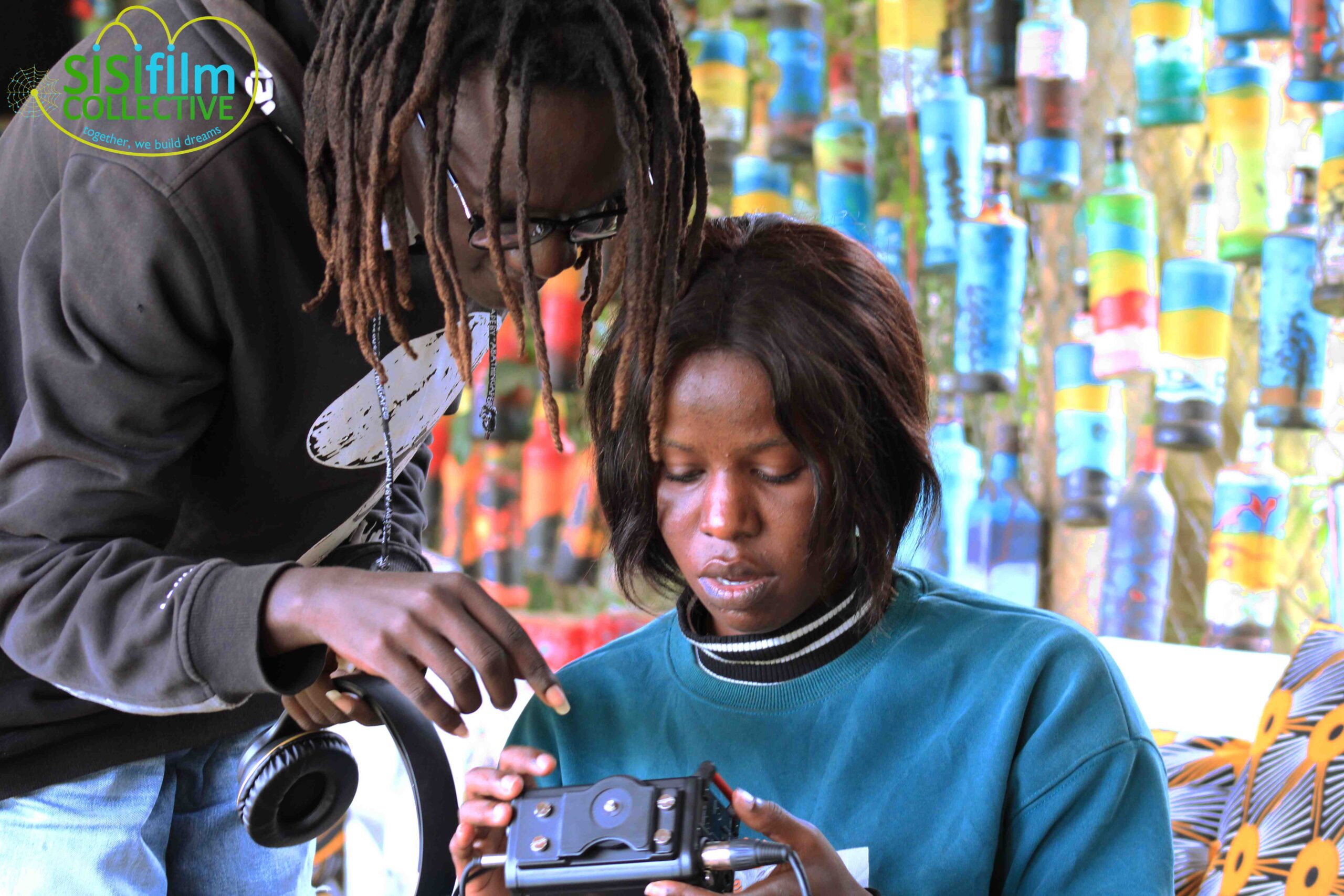 Two young African youths, a boy with dreads in a black jumper bending over showing something on a Tascam recorder to a girl with a wig dressed in a blue sweater while holding a Tascam.