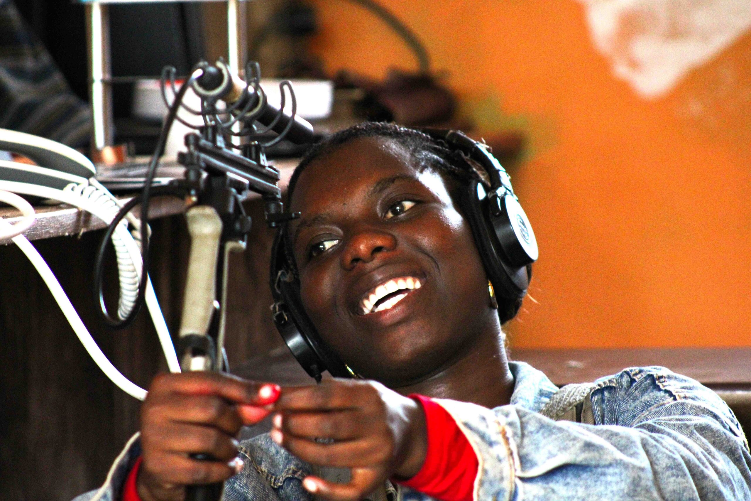 A closeup of a young African woman holding a professional boom pole and boom microphone, NTG2. She is smiling broadly and has headphones.