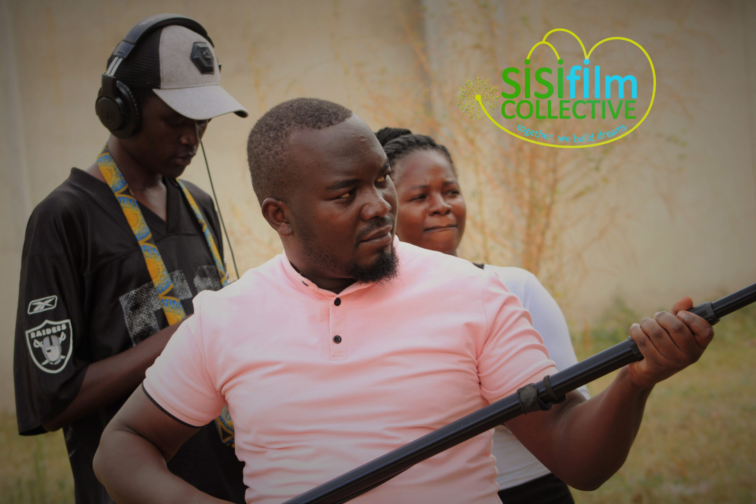 A young African man dressed in a pink shirt is carrying a boom as other young people are behind him.