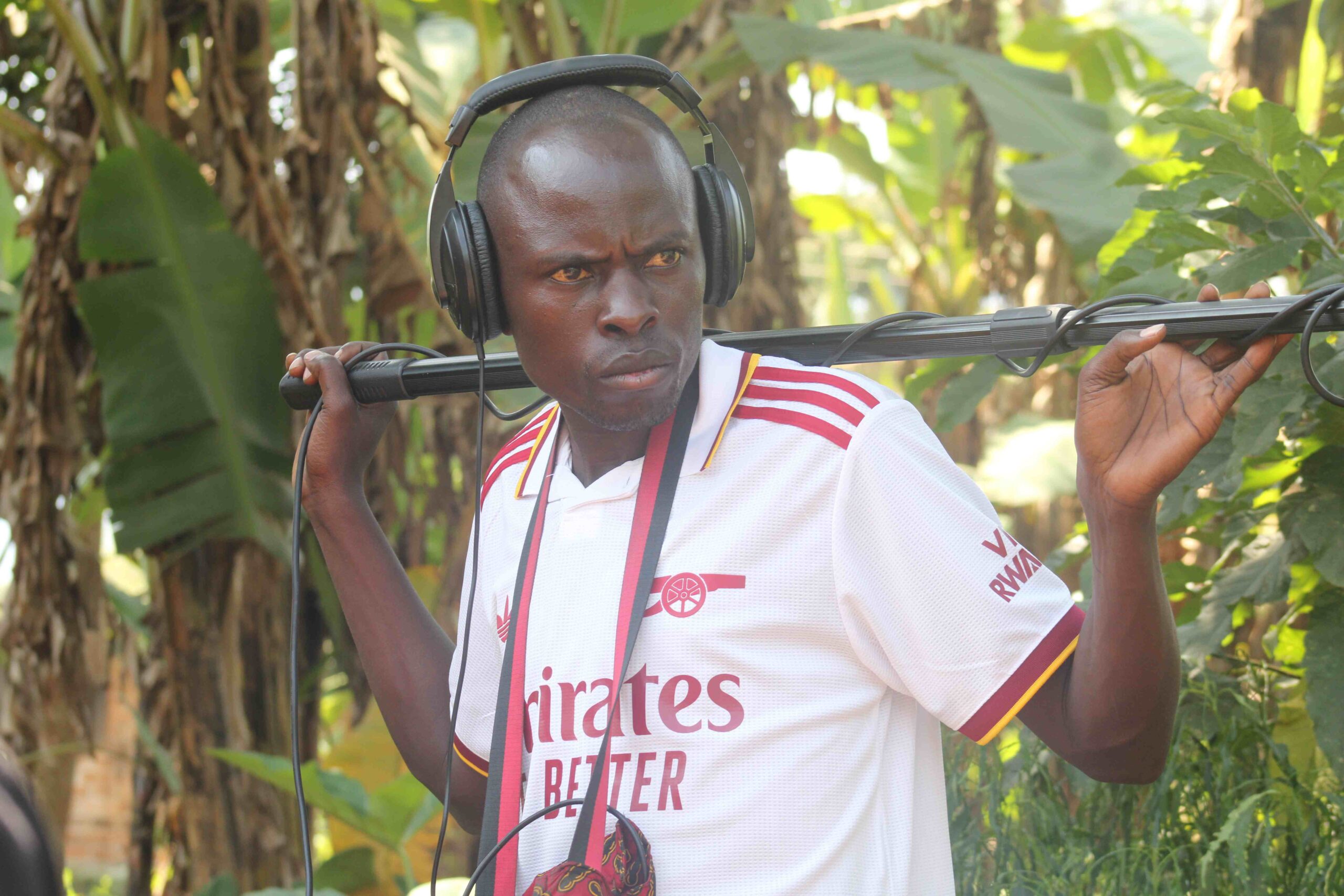 A young black African man carrying a boom and wearing headphones. He is dressed in a white fly emirates shirt with red strips.