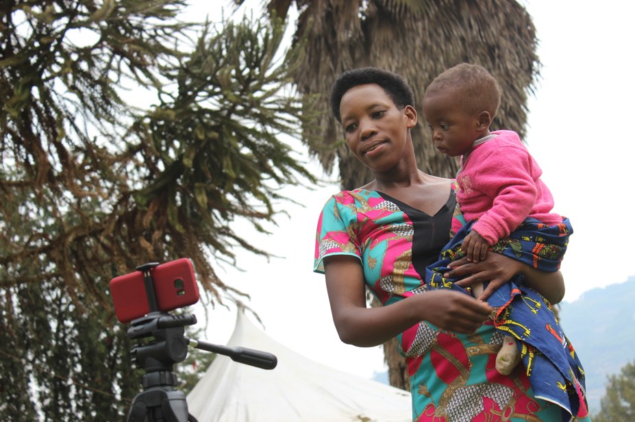 A lady stands carrying her baby. In front of her is a smartphone on a tripod.