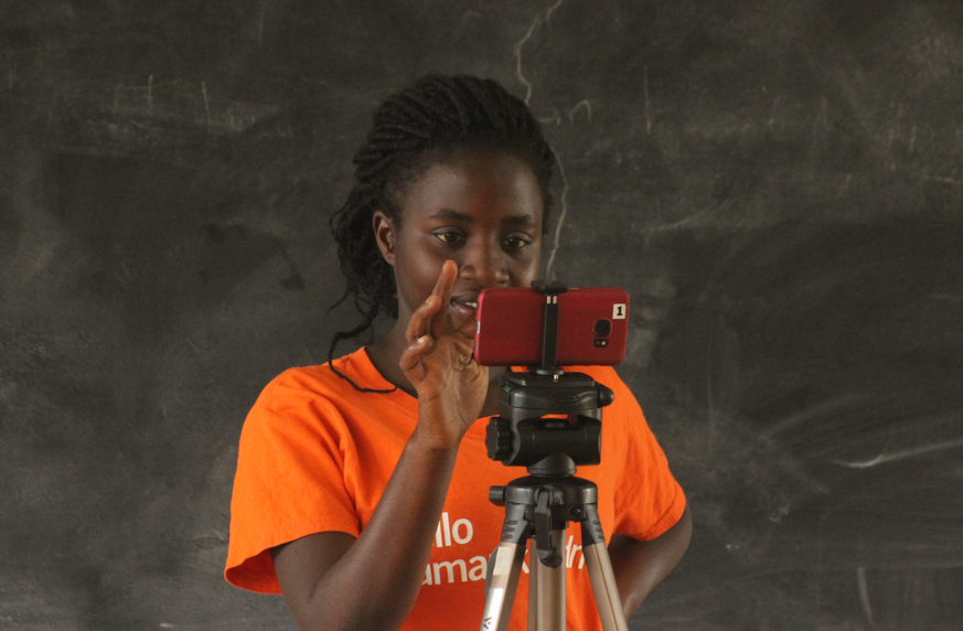 A young woman in an orange shirt adjusts a red smartphone on a tripod, focused on the screen.