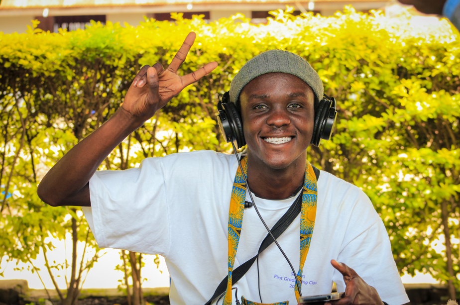 A young man dressed in a white shirt poses with a peace sign. He wears a head sock with headphones. He holds an African print bag around his neck.
