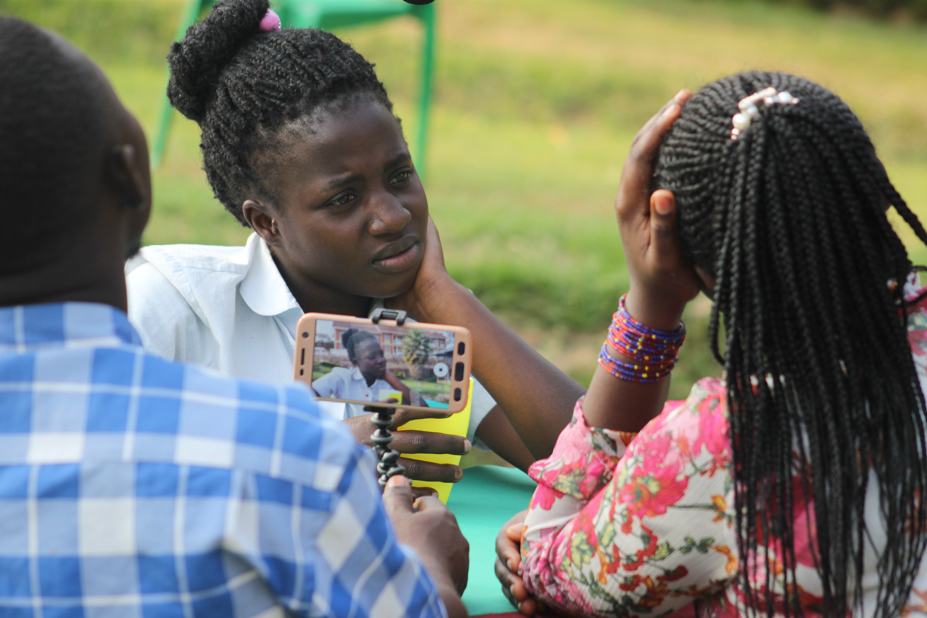 2 young people record an actor with a smart phone camera. The camera captures their backs as the actor is staring at something with her hand rested on her cheek