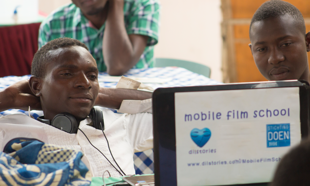 Two students during an editing class with another in the background. A laptop with a mobile film school poster sits before them.
