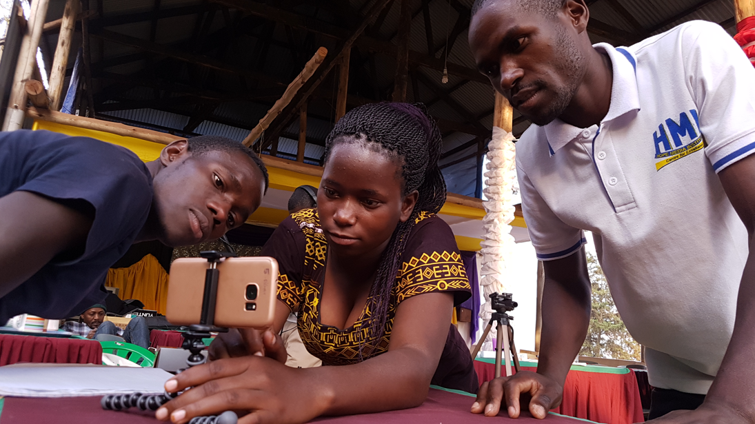 Three people lean over a table. concentrating on a smartphone mounted on a tripod.