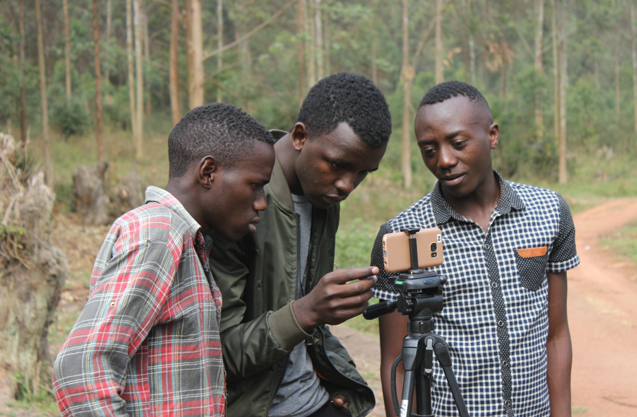 Three young people standing outdoors near a tripod-mounted smartphone. They are gathered closely together, looking at the phone screen as if reviewing footage or photos. Trees and a dirt path are visible in the background.