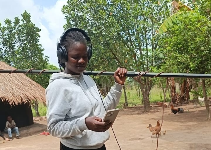 A young lady holds a boom pole on her shoulder. She wears headphones while she scrolls on a phone